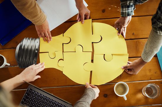 Glühlampe aus Puzzleteilen, die von mehreren Händen zusammengesetzt wird